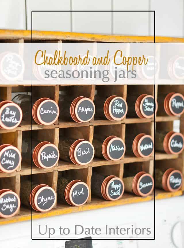 Glass spice jars with copper lids and round chalkboard labels lined up in a vintage yellow wooden soda crate on a counter