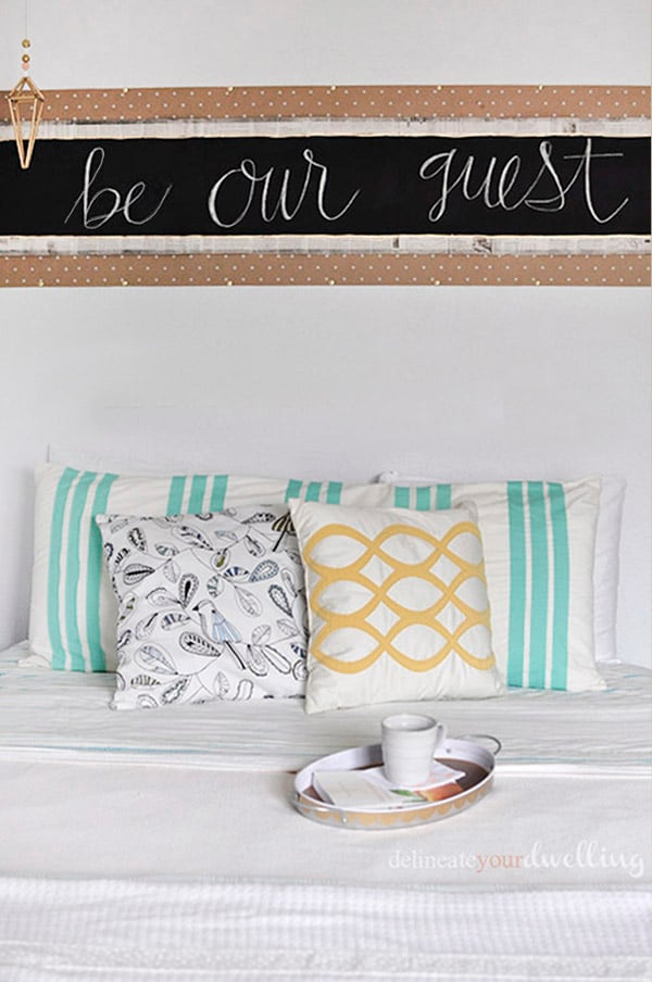 Chalkboard headboard reading be our guest in white chalk over newsprint and kraft paper above a bed with patterned pillows