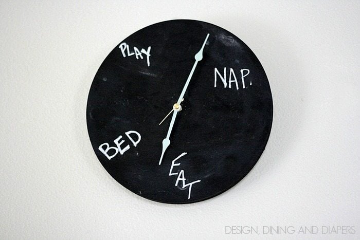 Round black chalkboard clock on a white wall with play, nap, eat and bed hand-lettered in chalk and pale aqua clock hands
