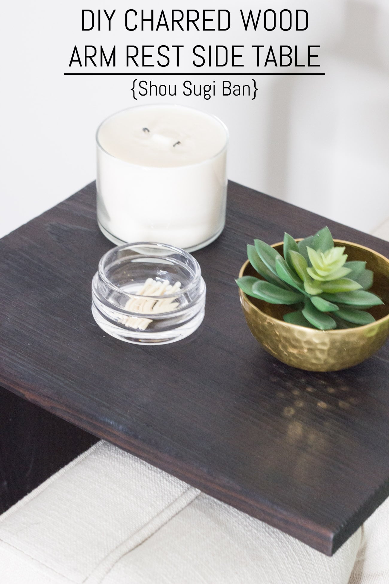 Charred black wood arm rest table over a cream sofa arm holding a white candle, a glass dish and a gold bowl with a succulent