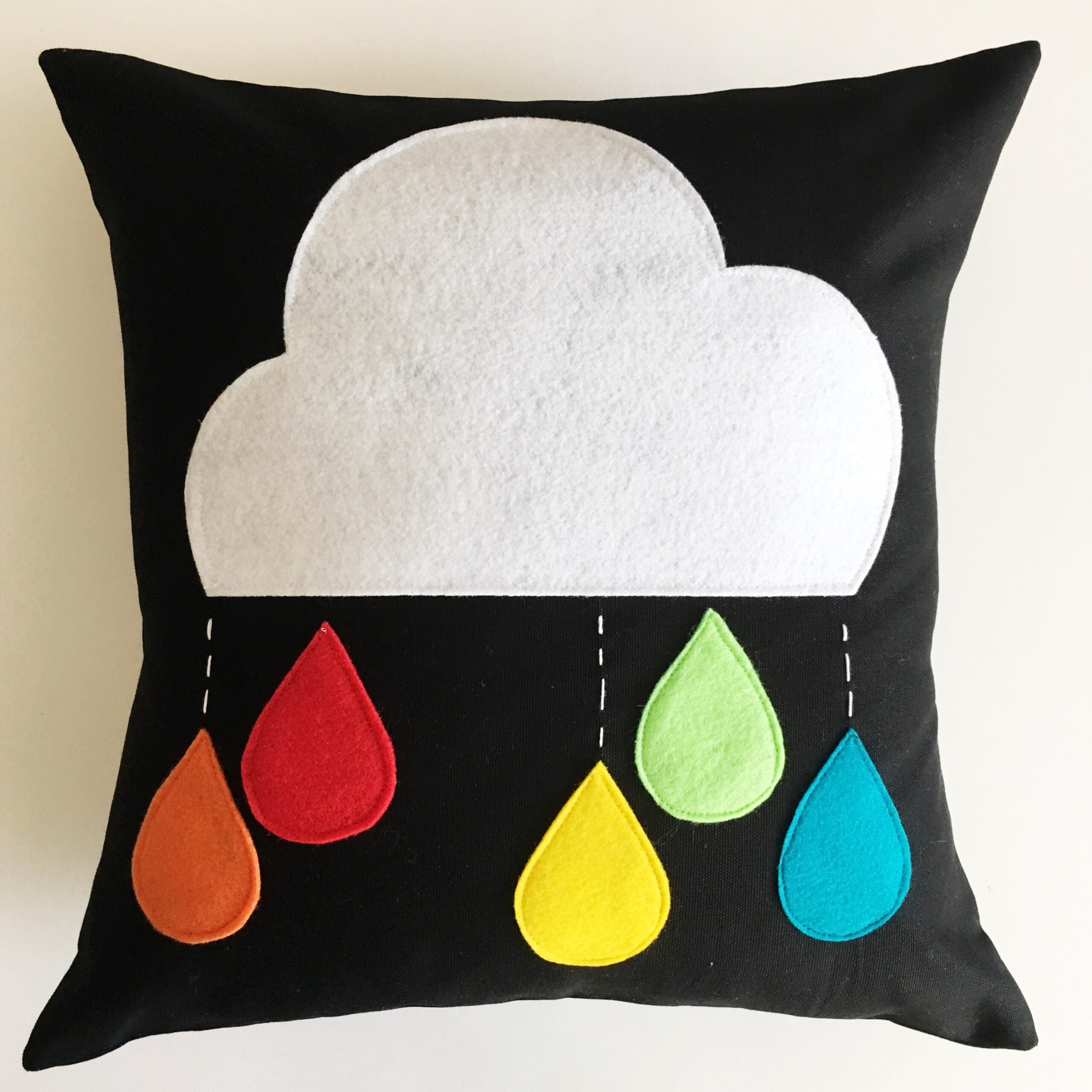 Black throw pillow with a large white felt cloud and five rainbow felt raindrops in red, orange, yellow, green and blue
