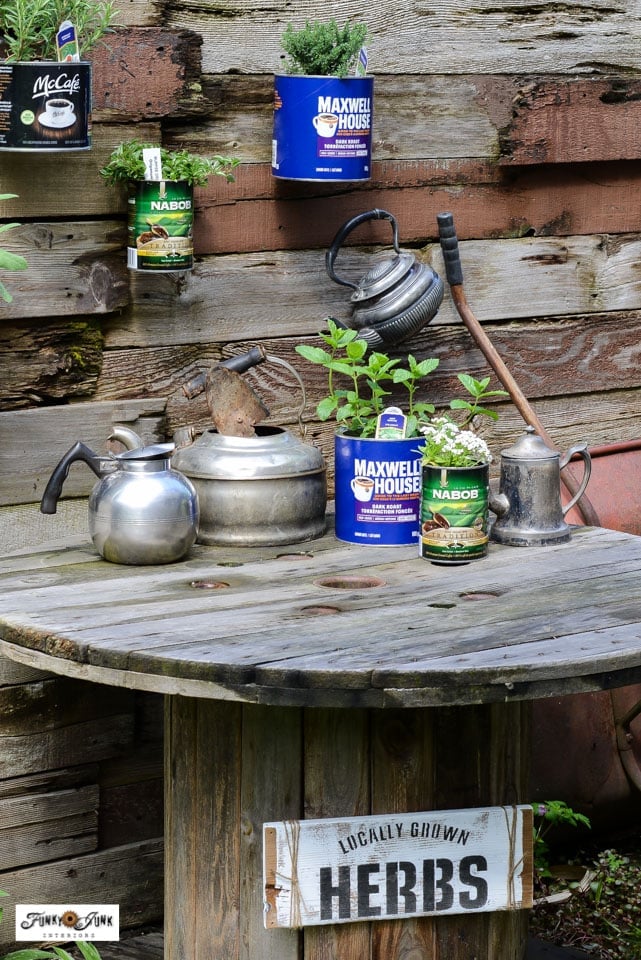Coffee Can Herb Garden on a Fence | Tin Can Idea Coffee can herb planters with their colorful labels still on, mounted on a wooden fence above a spool table and herbs sign