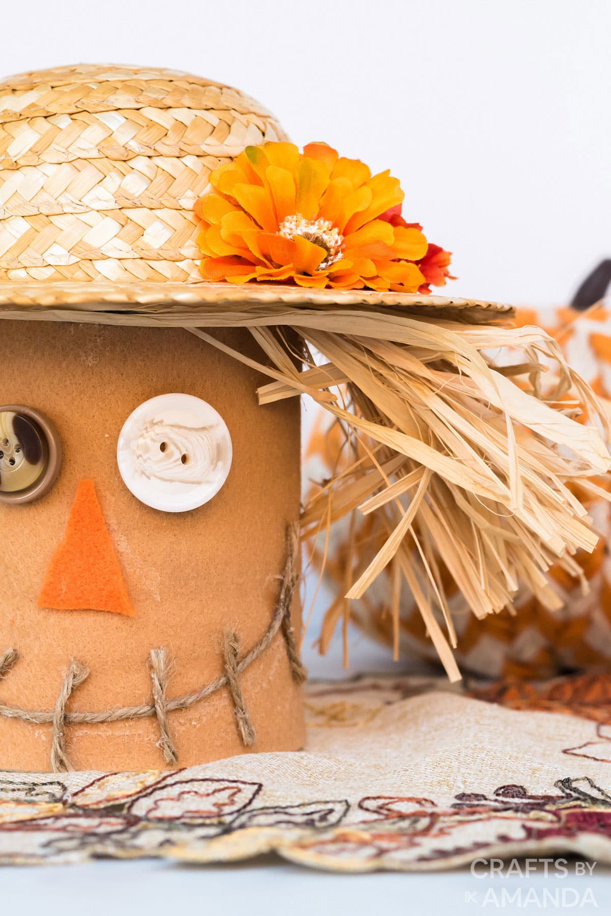 Coffee can scarecrow covered in tan felt with a straw hat, button eyes, an orange felt nose and a stitched twine smile