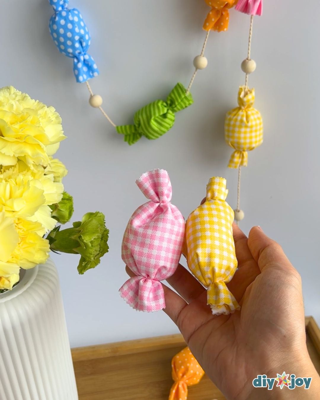 Hand holding pink gingham and yellow gingham fabric candies beside yellow carnations, with the hanging garland behind