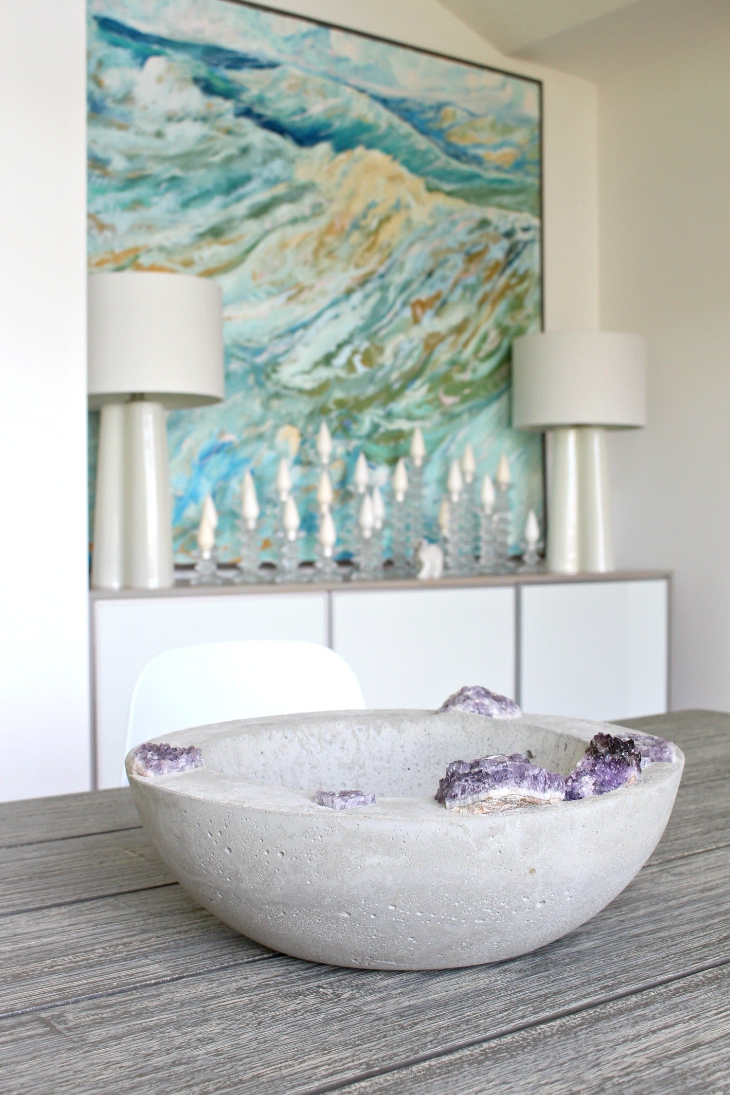 Concrete Bowl with Amethyst | Geode Bowl DIY Wide gray concrete bowl with purple amethyst chunks set into the rim on a wood table in a white room with a blue painting