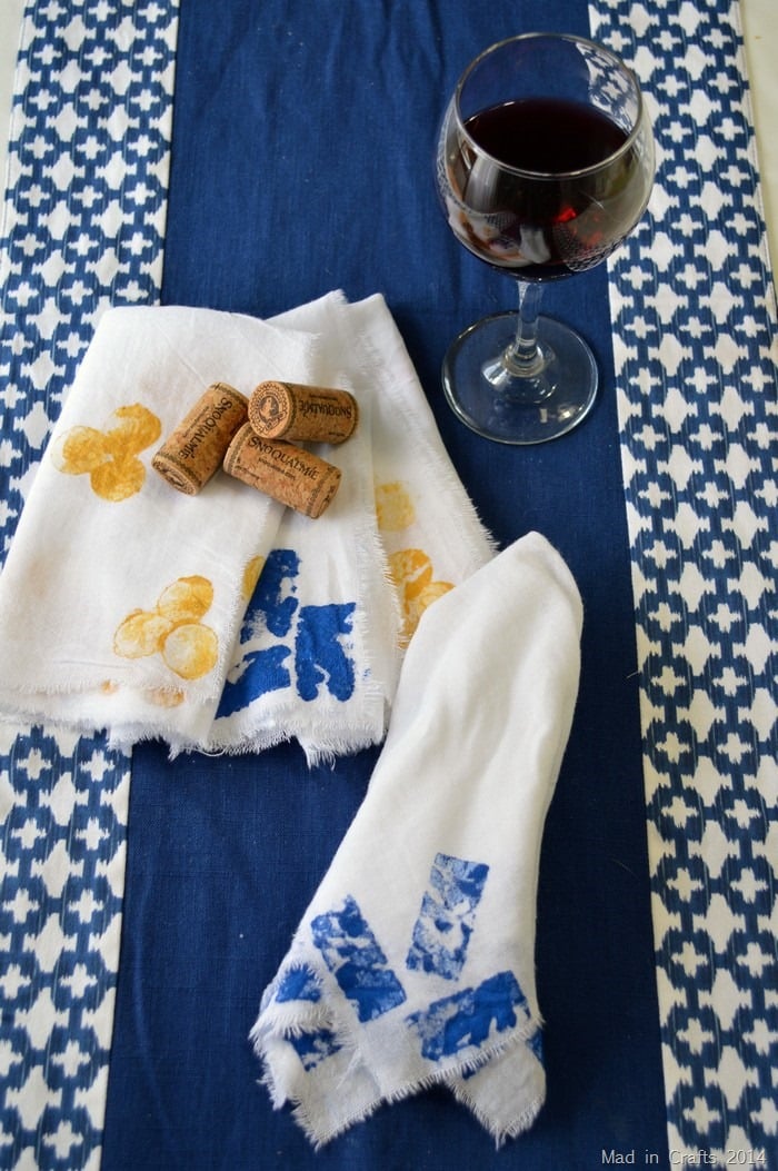 Cork stamped raw edge napkins with yellow dots and blue rectangles on white cloth, beside wine corks and a glass of red wine