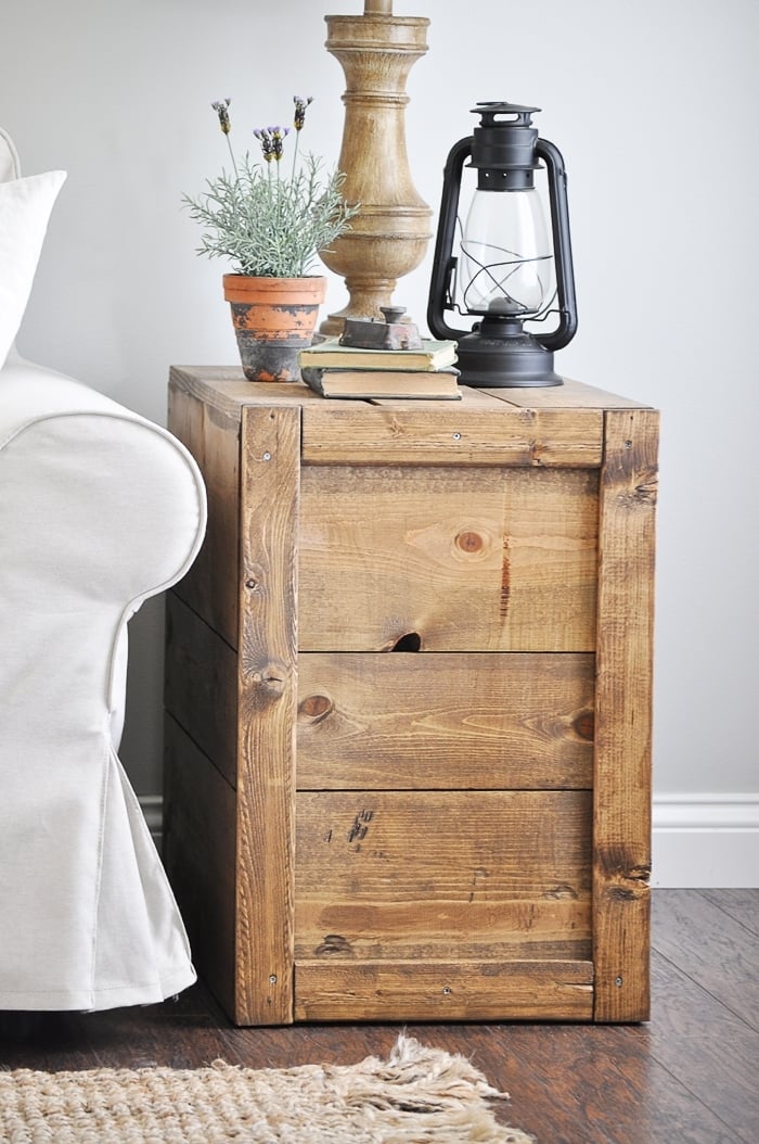 Rustic stained wood crate style side table beside a white slipcovered chair with a lamp, a lantern and a potted lavender