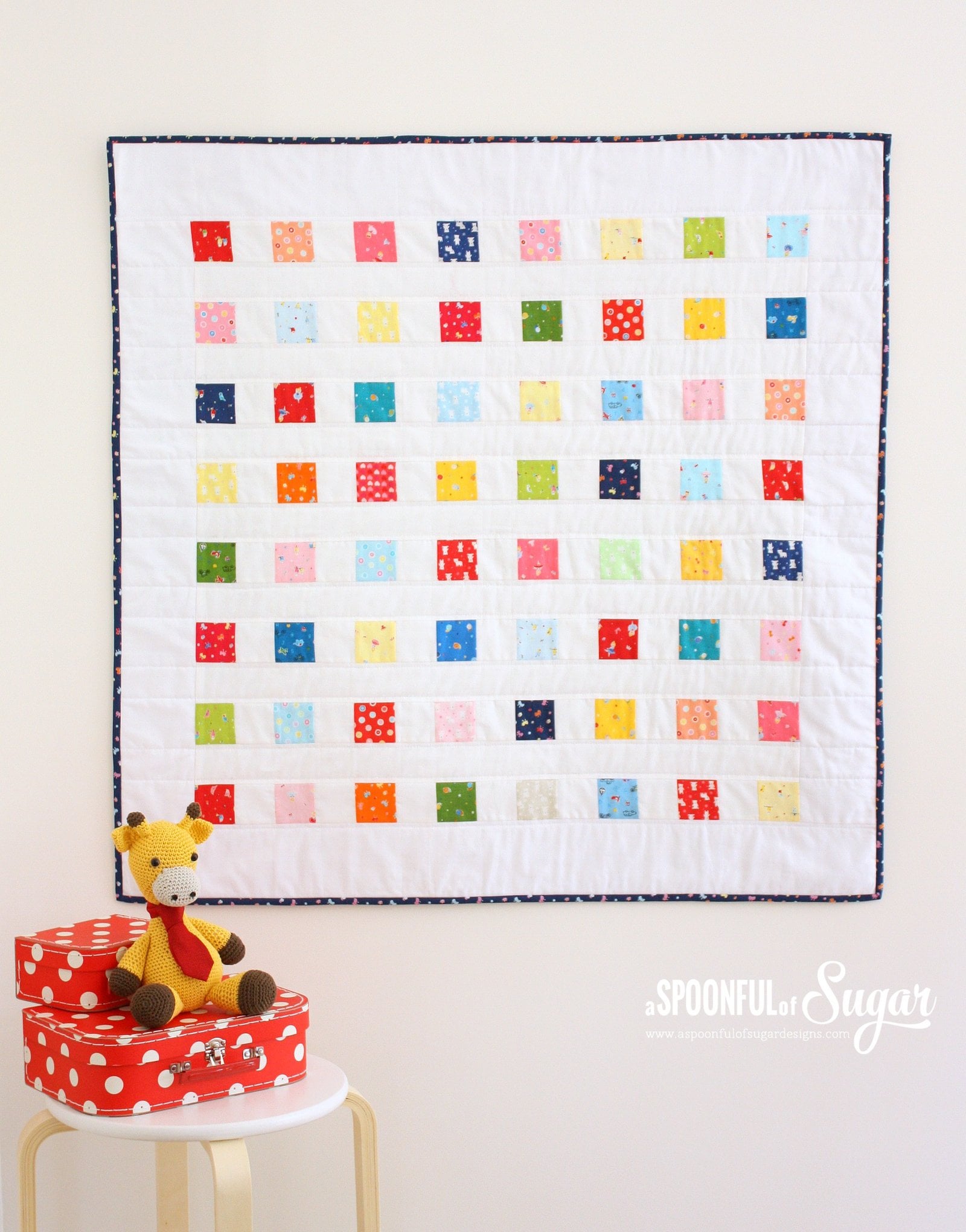 Square white baby quilt with rows of small colorful patchwork squares hanging above a red polka dot box