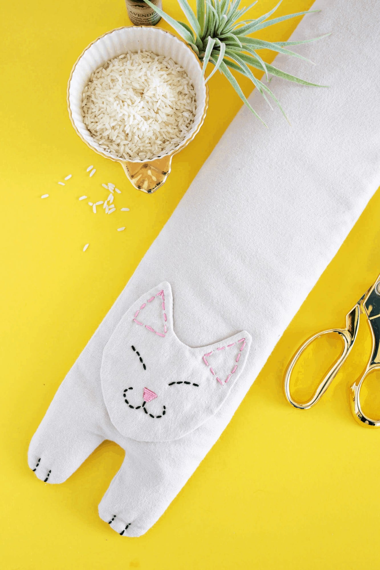 Kitty Rice Heating Pad Sewing Gift White cat shaped rice heating pad with an embroidered sleepy face, ears and paws on a yellow table with a bowl of rice