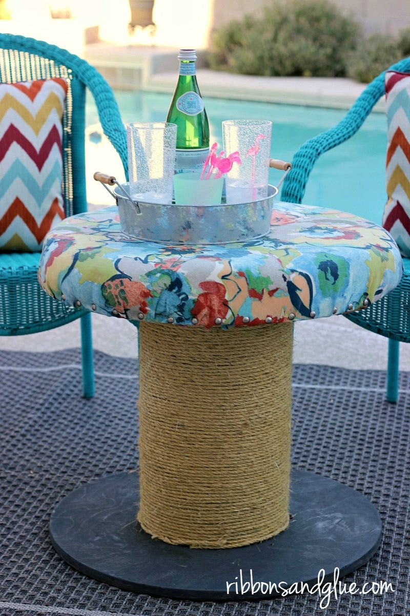 Wooden cable spool table with a rope wrapped base and a padded floral fabric top, holding drinks by a pool with blue chairs