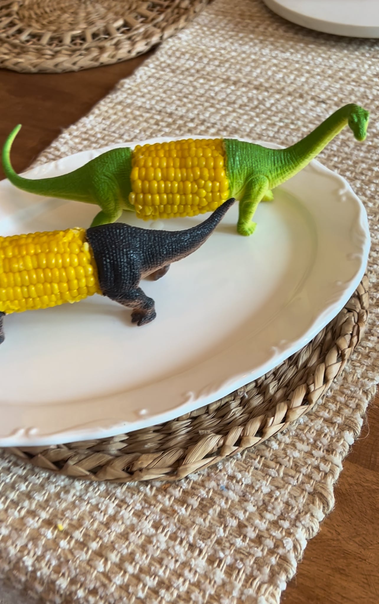 Green and dark brown toy dinosaurs cut in half and holding corn on the cob as a body on a white plate over a woven placemat