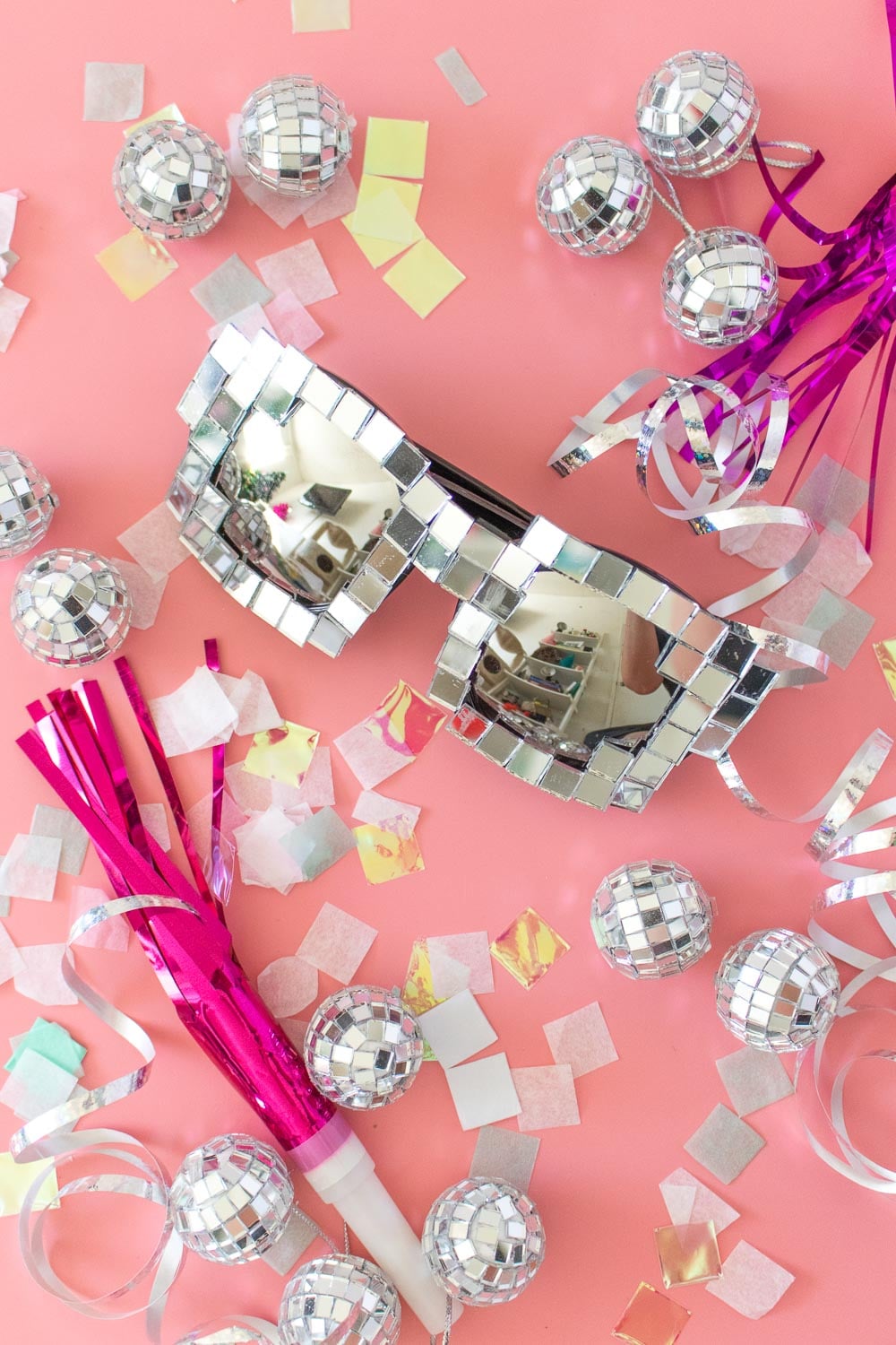 Sunglasses covered in tiny mirror squares on a pink background with mini disco balls, confetti and pink noisemakers