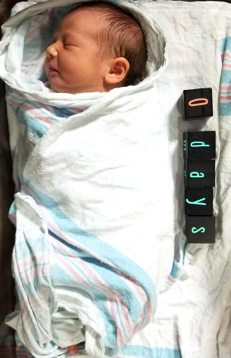 Newborn swaddled in a striped hospital blanket beside a stack of black chalkboard milestone blocks spelling 0 days in vinyl
