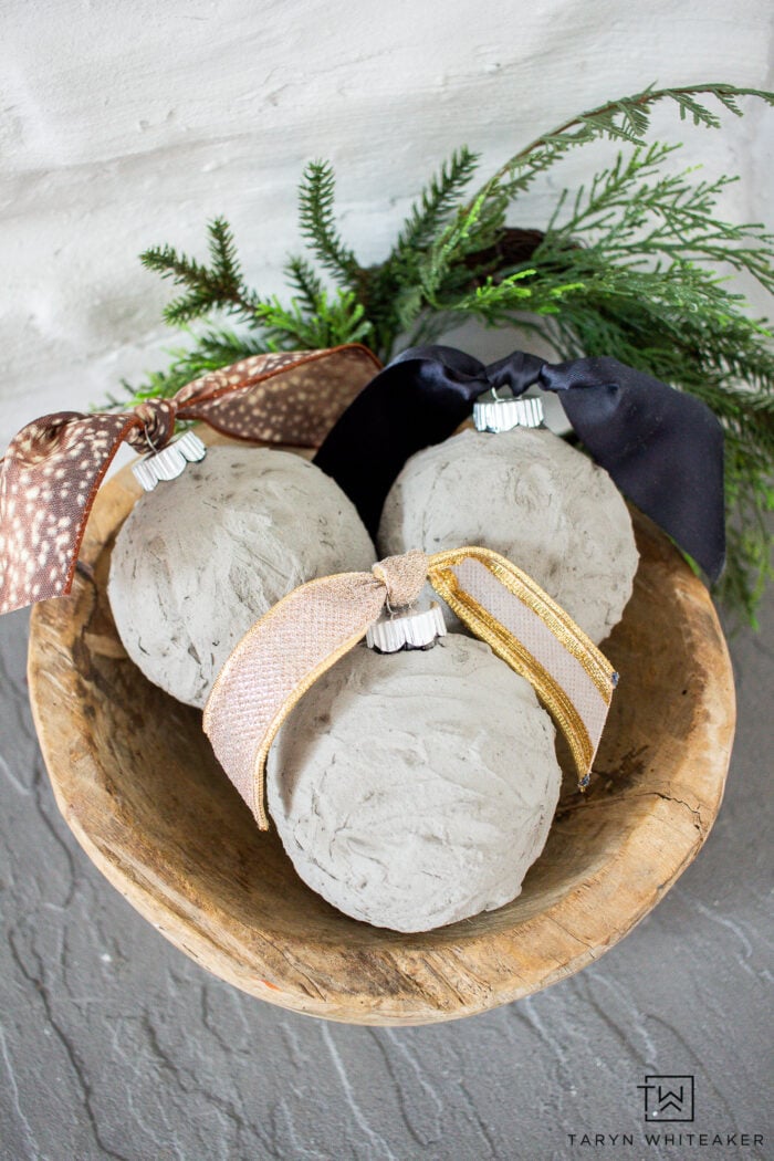DIY Concrete Christmas Ornaments | Industrial Chic Three textured gray concrete ball ornaments with brown dotted, black and gold ribbons in a wood bowl with greenery