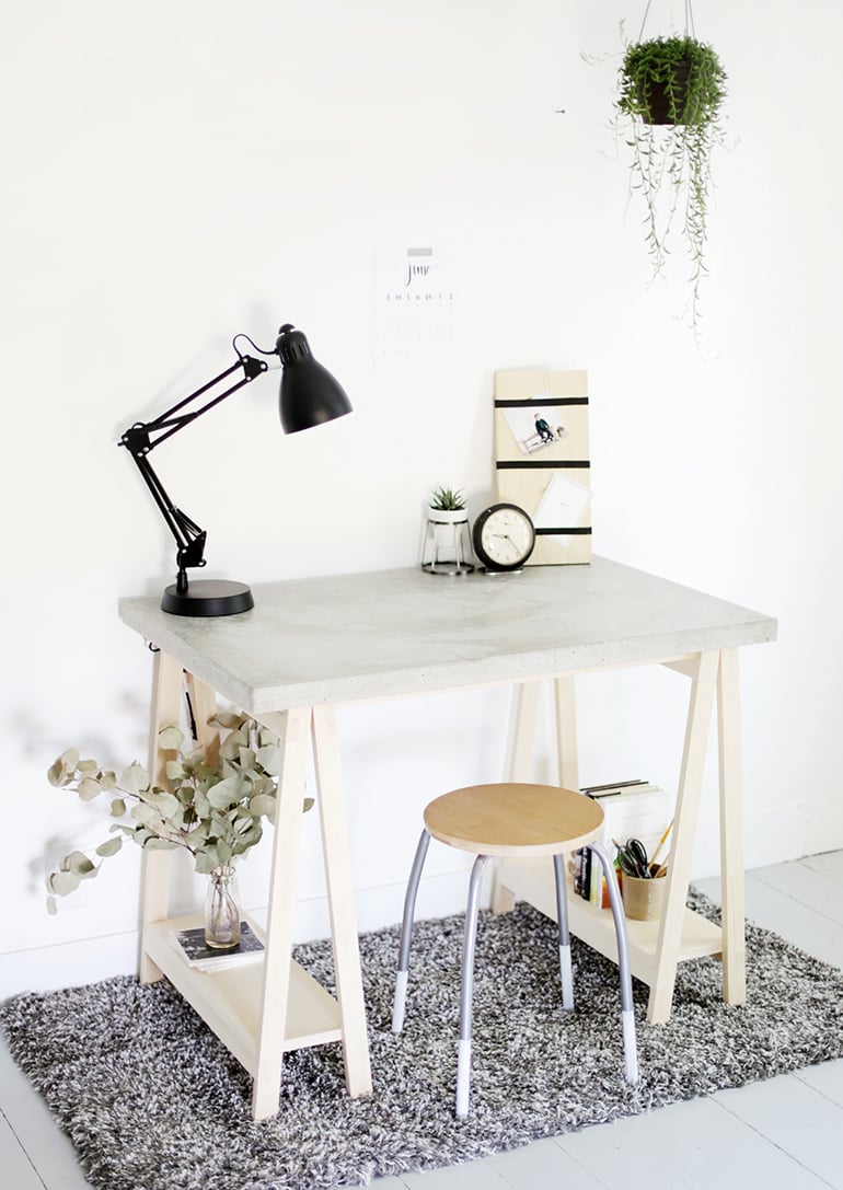 DIY Concrete Desk | Sawhorse Leg Workspace White concrete topped desk on light wood sawhorse legs with a black lamp, a stool and a hanging plant in a white room