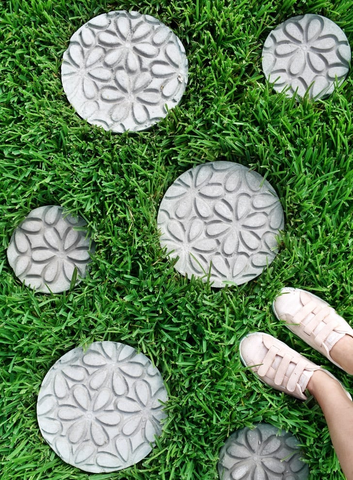 Embossed Stepping Stones | Doormat Stamp DIY Round gray concrete stepping stones with embossed floral patterns set in green grass beside a pair of white sneakers