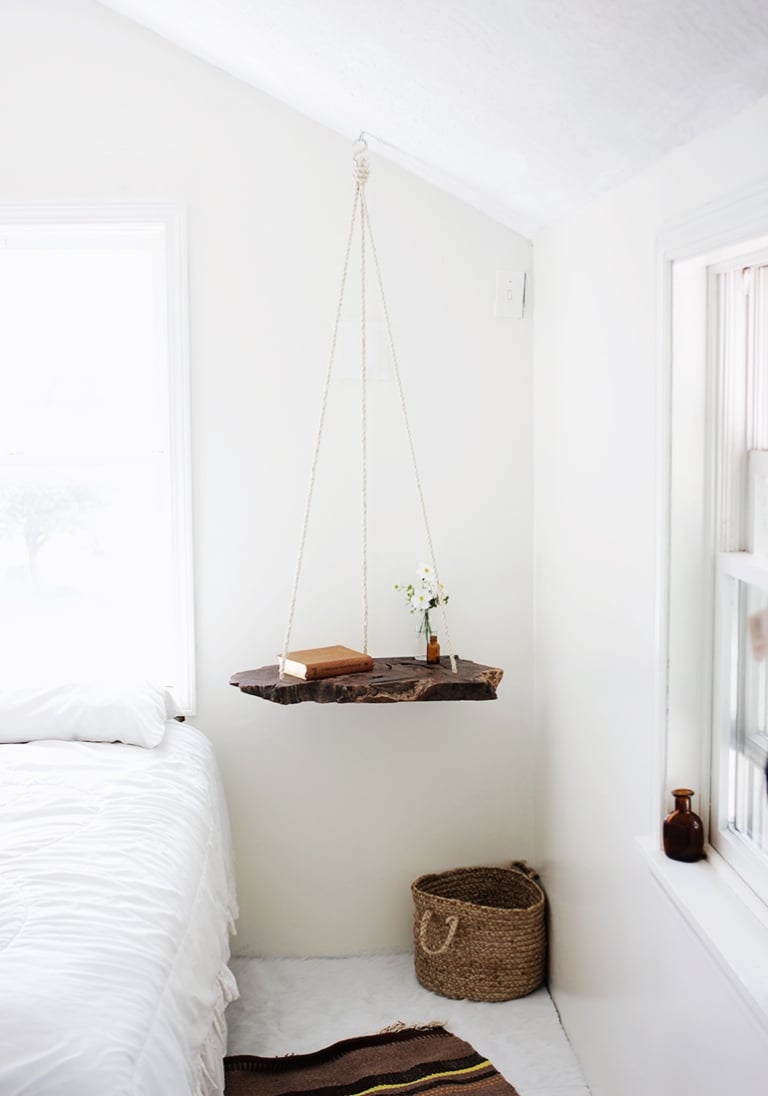 Live edge wood slab nightstand hanging from thick rope beside a white bed, holding a small vase of white flowers and books