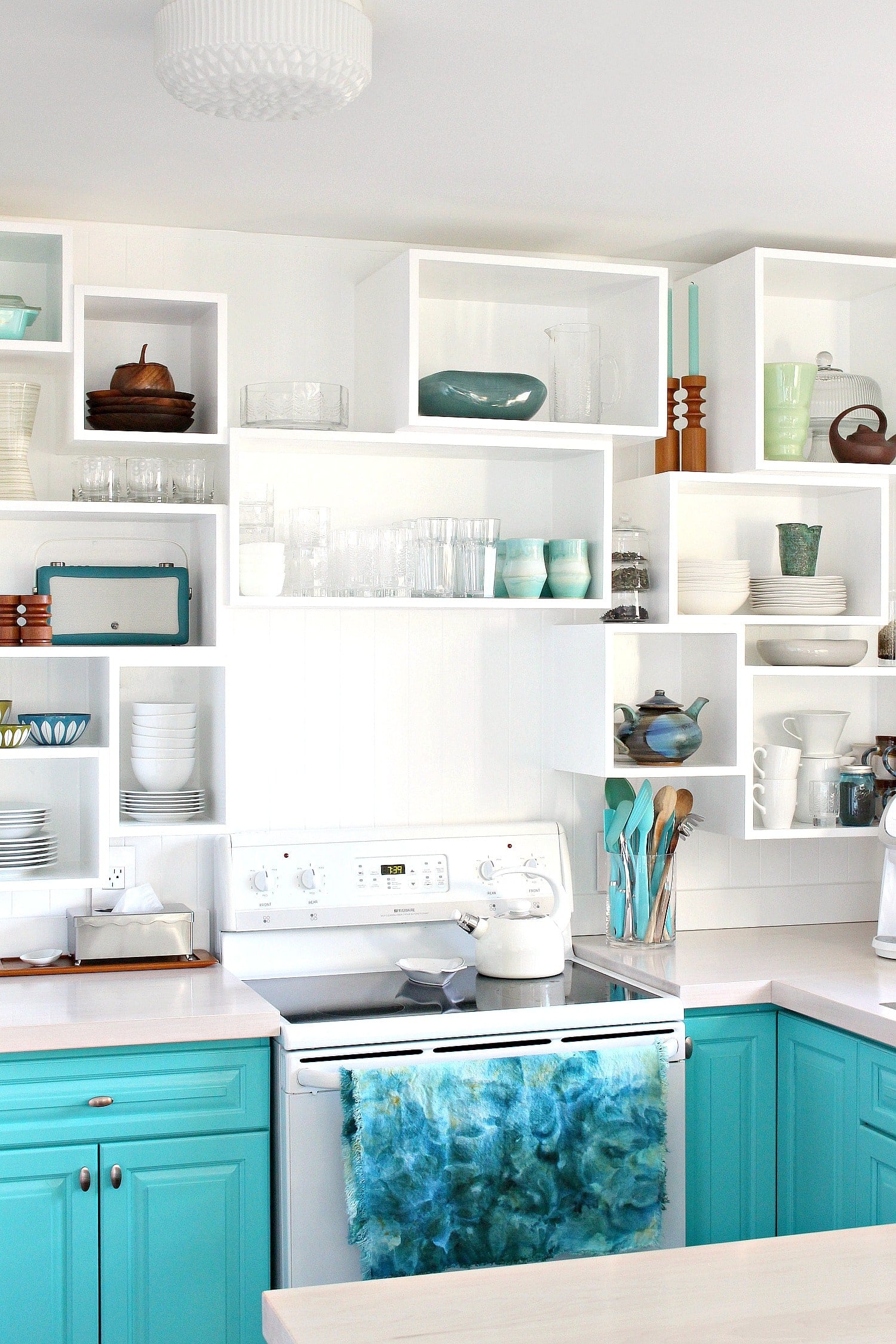 White kitchen wall of open wood cubbies in staggered sizes holding dishes, glasses and teal accents above a stove
