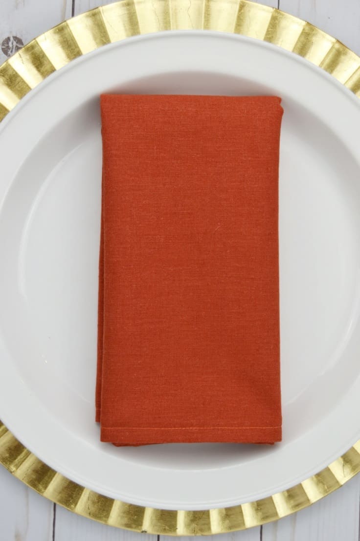 Easy DIY Linen Cloth Napkins to Sew Folded burnt orange linen cloth napkin on a white dinner plate set on a gold ruffled charger, white wood below