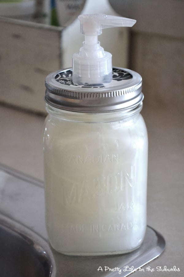 Mason jar filled with creamy white hand soap with a patterned paper covered lid and a clear pump beside a kitchen sink