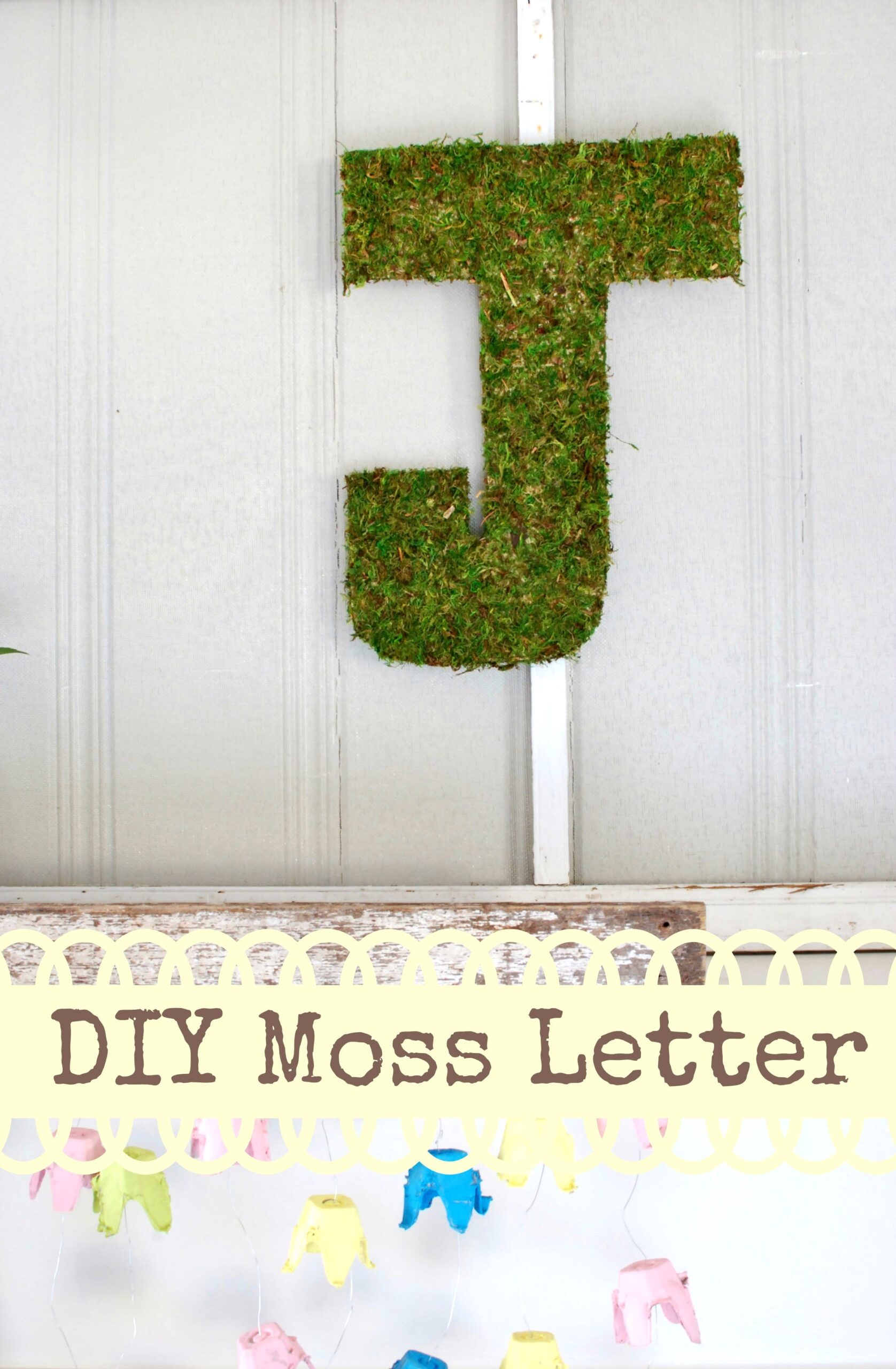 Green moss covered letter J hanging on a pale paneled wall above a pastel painted egg carton garland