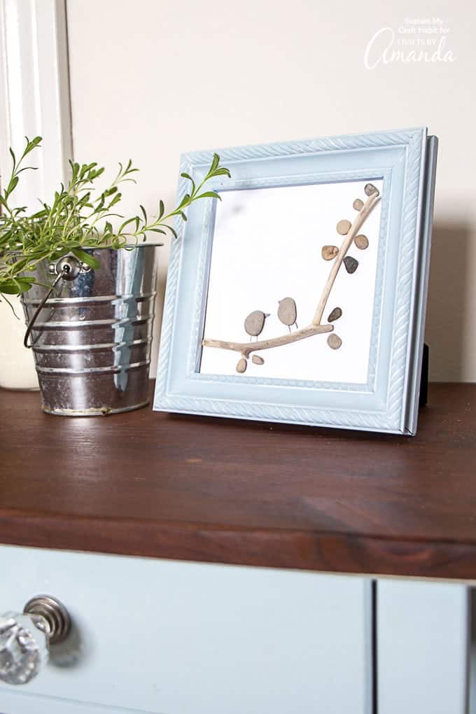 Framed pebble art of two small stone birds perched on a driftwood twig with pebble leaves, set on a dresser beside herbs