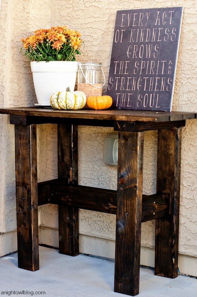 DIY Porch Table | DIY Patio Furniture Idea Dark stained wood porch table holding orange mums and small pumpkins in front of a chalkboard sign on a stucco wall