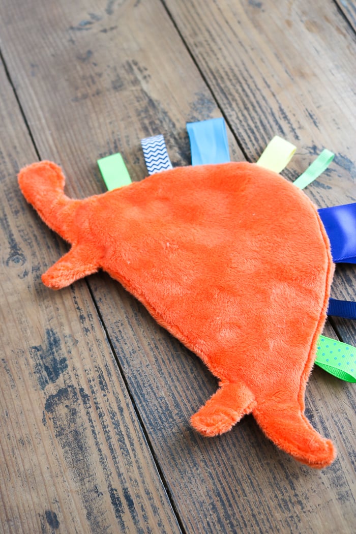 Orange minky dinosaur shaped taggie blanket with blue, green, yellow and chevron ribbon tags along its back on gray wood