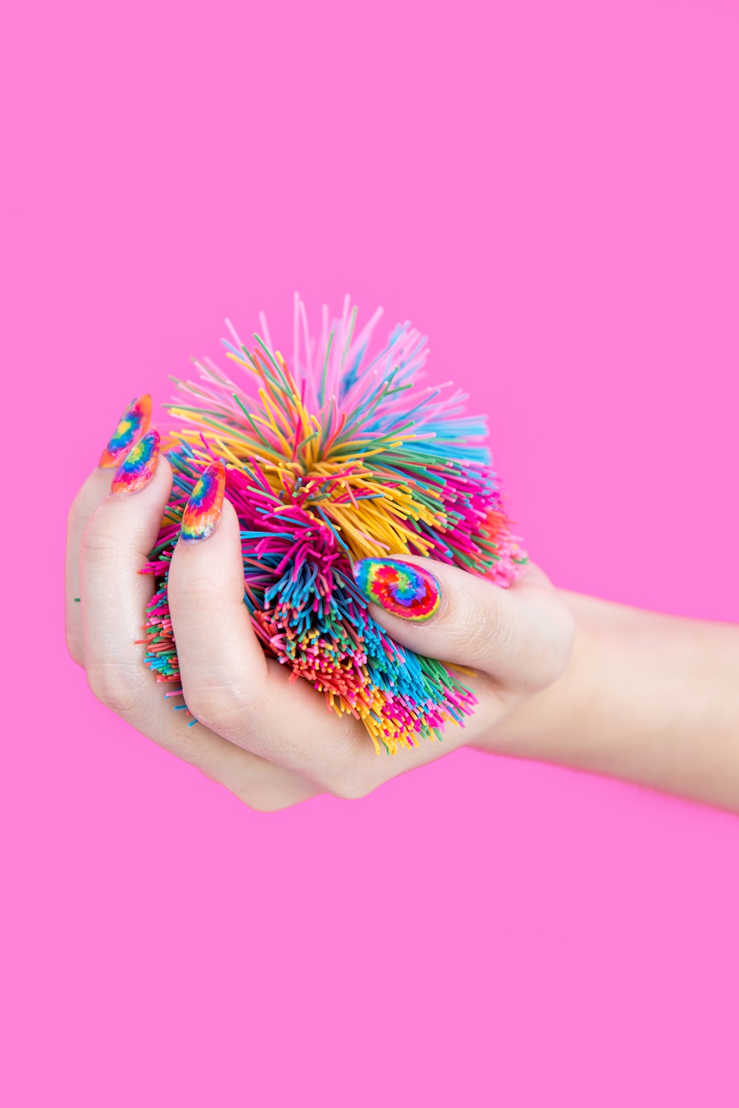 DIY Tie-Dye Nail Wraps | Quick Nail Art Idea Hand with rainbow tie-dye nail wraps in spiral swirls, squeezing a colorful stringy toy ball against a hot pink background
