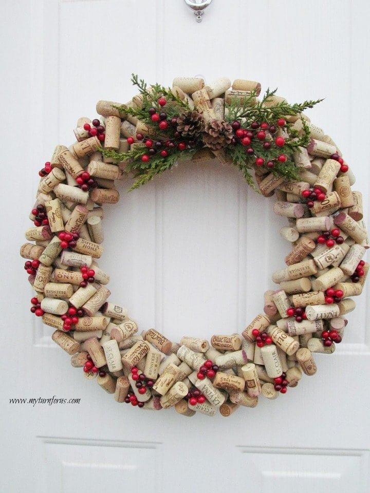 DIY wine cork wreath of layered overlapping corks with evergreen sprigs, pinecones and red berries on a white door