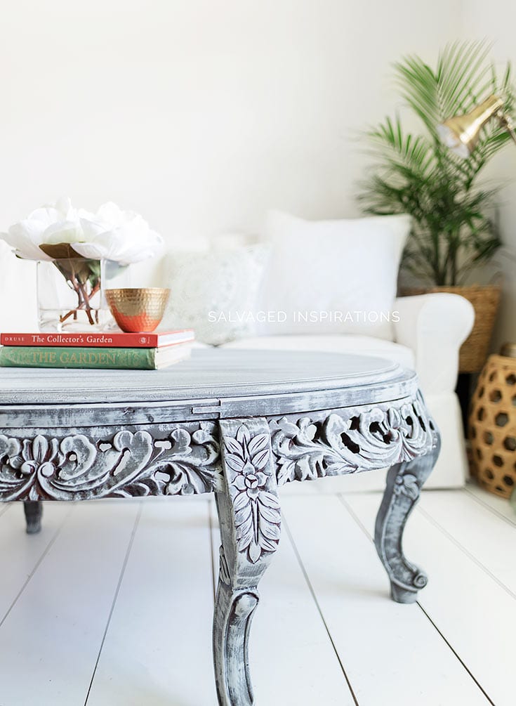 Oval carved wood coffee table dry brushed in light and dark gray, styled with books and white flowers in a white room