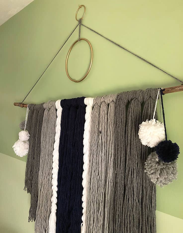 Gray, black and white yarn wall hanging with braided strands and white pom poms on a stick with a gold ring on a green wall