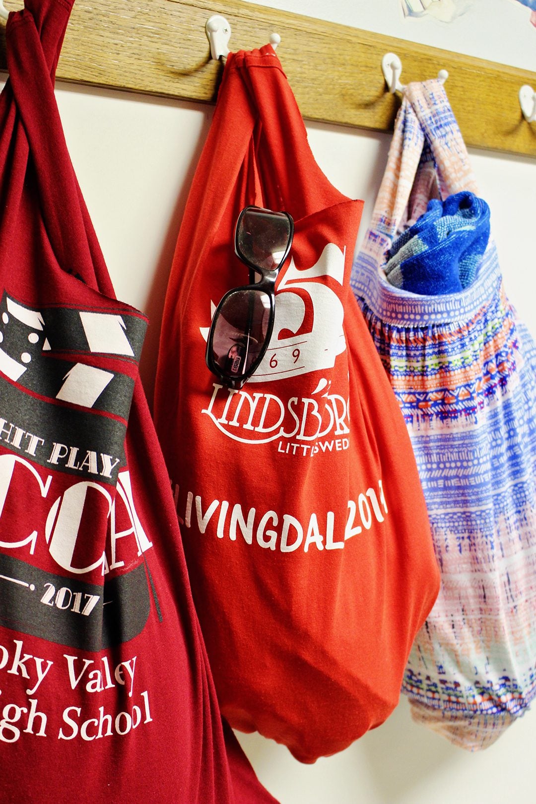 Three upcycled T-shirt tote bags on wall hooks, two red graphic tees with sunglasses and one patterned knit with a towel