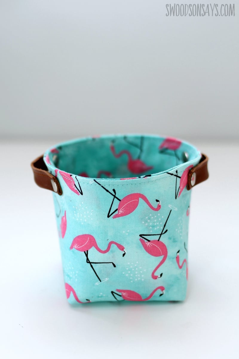 Small round fabric basket in aqua flamingo print cotton with brown leather tab handles on a white table
