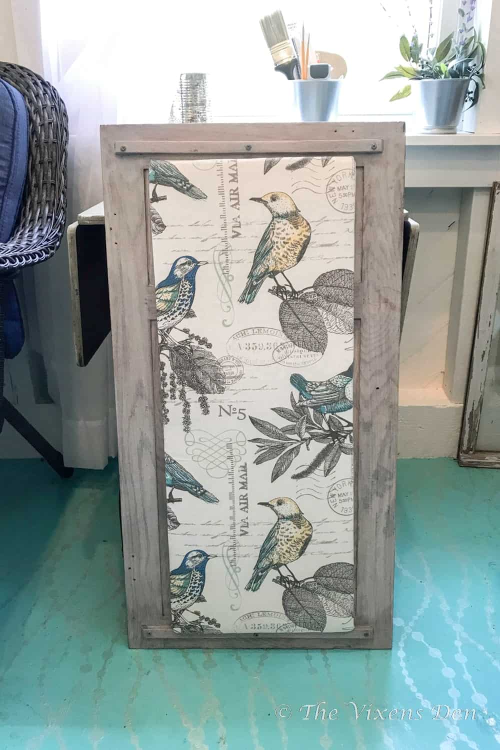 Gray wood cabinet door with a fabric covered bulletin board in a cream bird and botanical print, crossed with tacks and twine