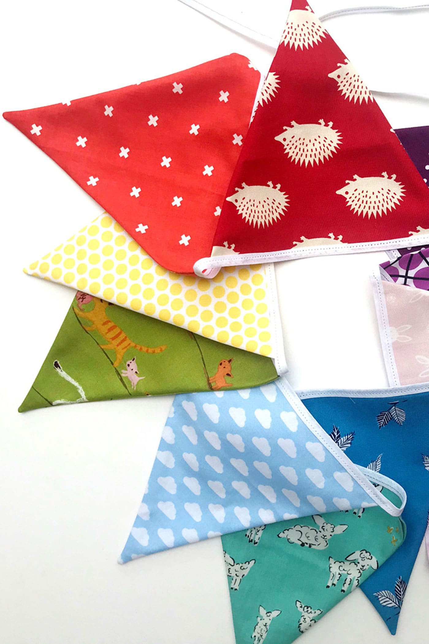 Fabric bunting flags in red with white crosses, hedgehogs, yellow dots, blue clouds and teal deer prints on white binding