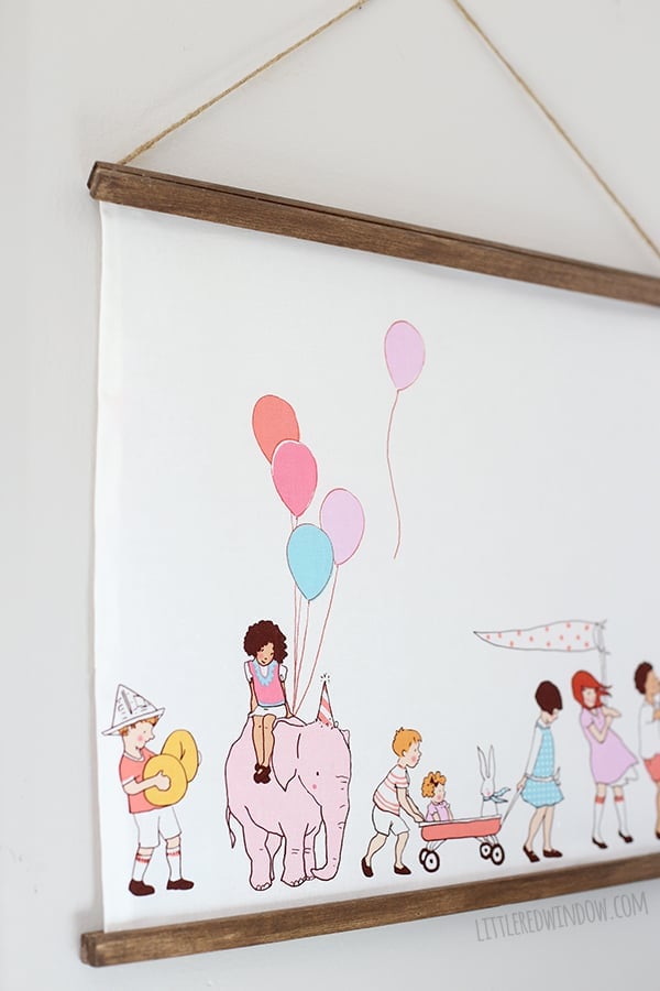 Fabric wall hanging on walnut wood rods printed with illustrated children in a parade with balloons and a pink elephant