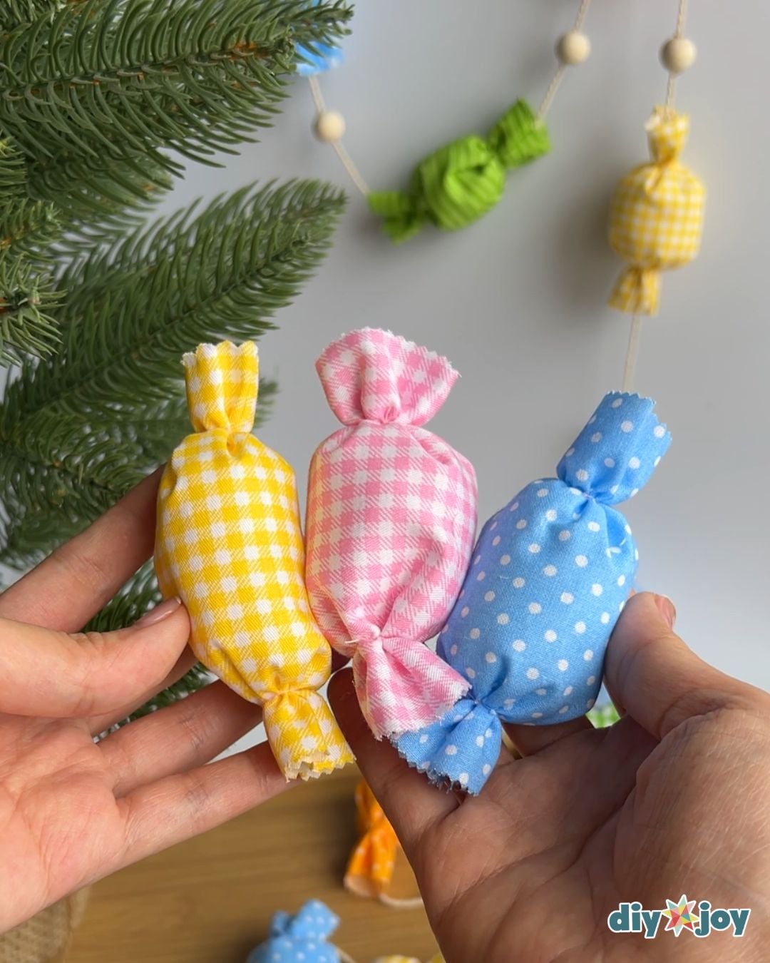 Two hands holding yellow gingham, pink gingham and blue polka dot fabric candies in front of a pine branch