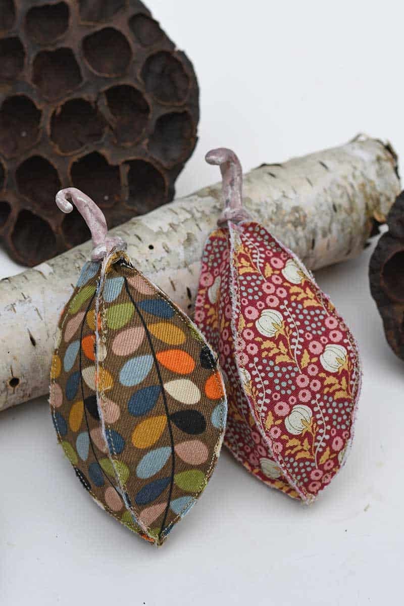 Two stuffed fabric seed pods in a mod leaf print and a red floral with curled clay stalks beside a birch log and lotus pod