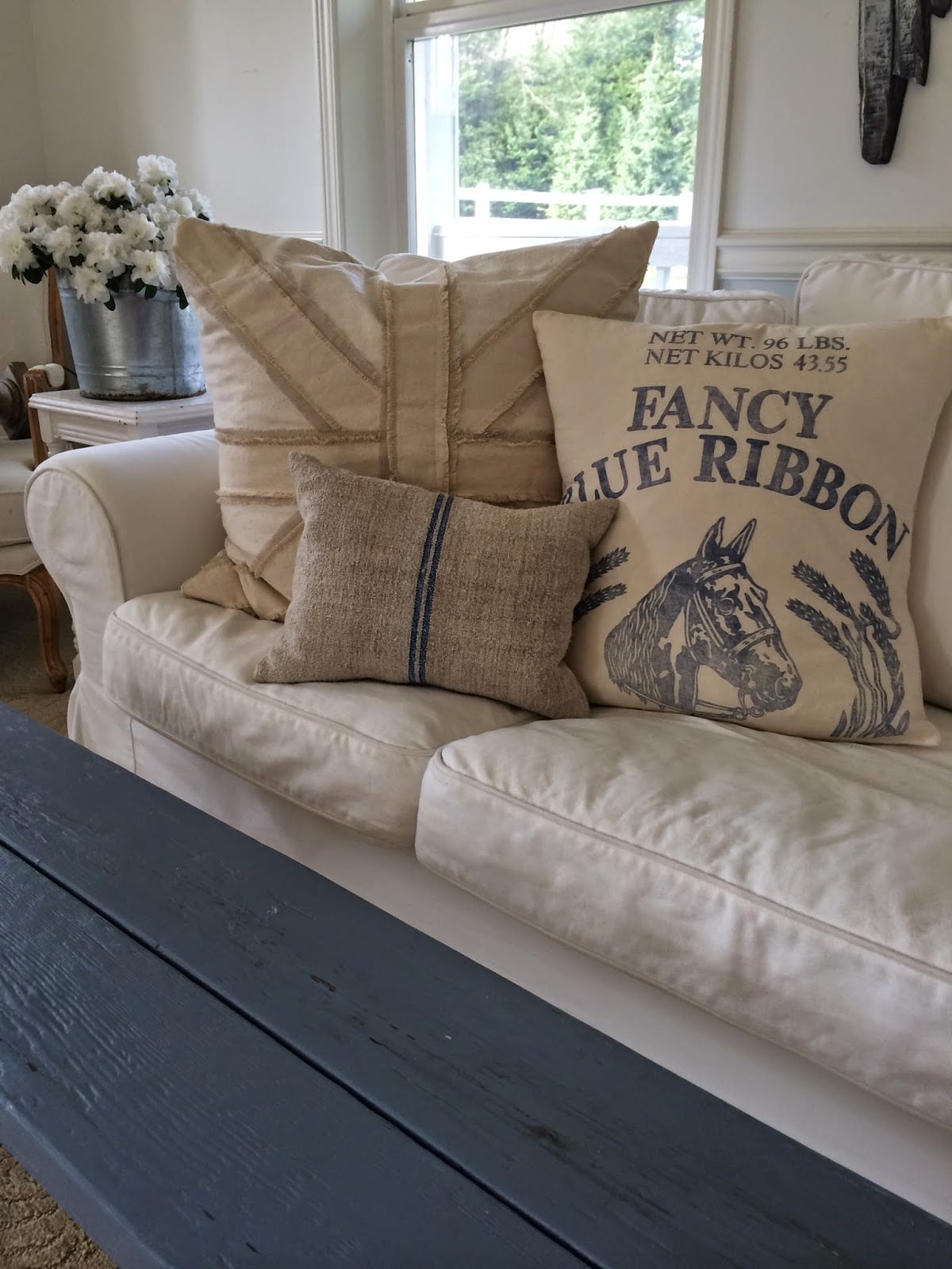 Farmhouse style porch with burlap, grain sack and horse print pillows on a white sofa beside a bucket of white daisies