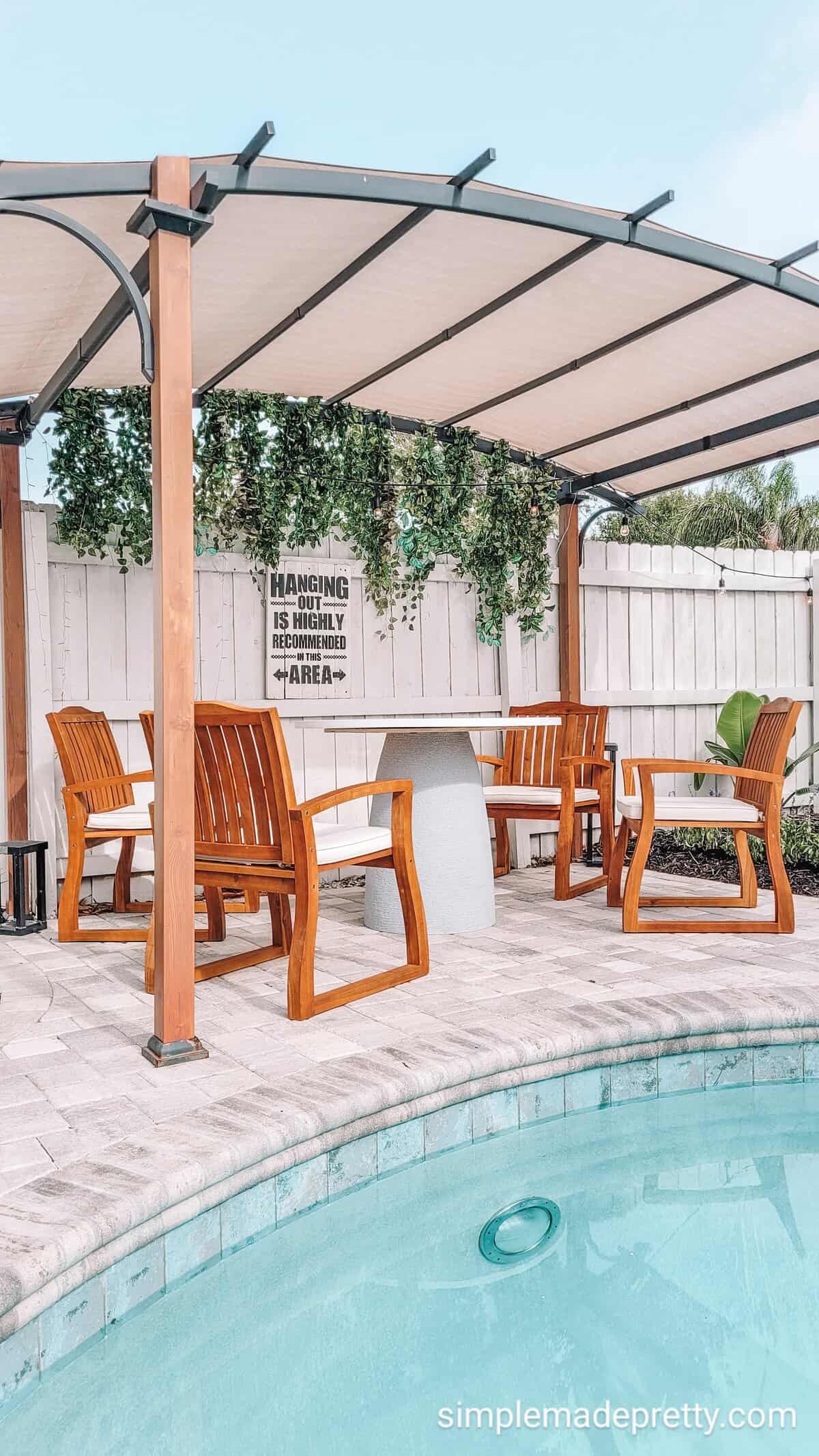 Faux Concrete Dining Table | DIY Patio Furniture Idea Round faux concrete outdoor dining table with wood chairs beside a pool under a curved pergola on a paver patio