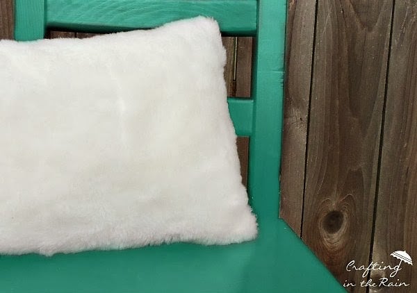 Faux Fur Pillow | Home Sewing Project Fluffy white faux fur pillow resting on a teal painted wooden chair seat against a rustic barn wood wall