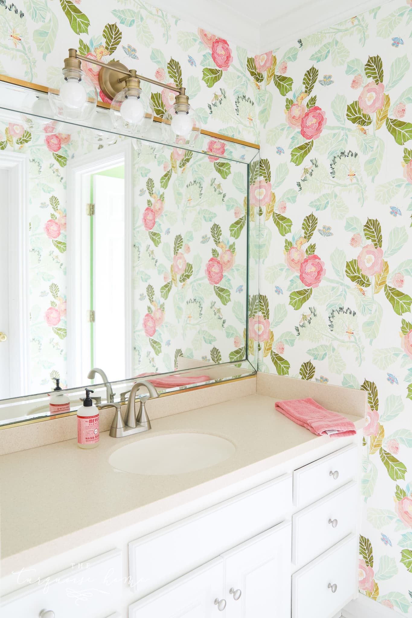 Bathroom wallpapered in pink and green watercolor florals with a wide mirror, brass light and white vanity