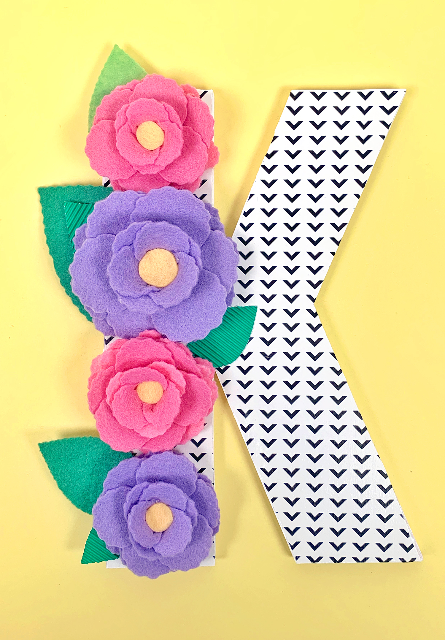 Black and white chevron patterned letter K with a column of pink and purple felt flowers and green leaves on yellow