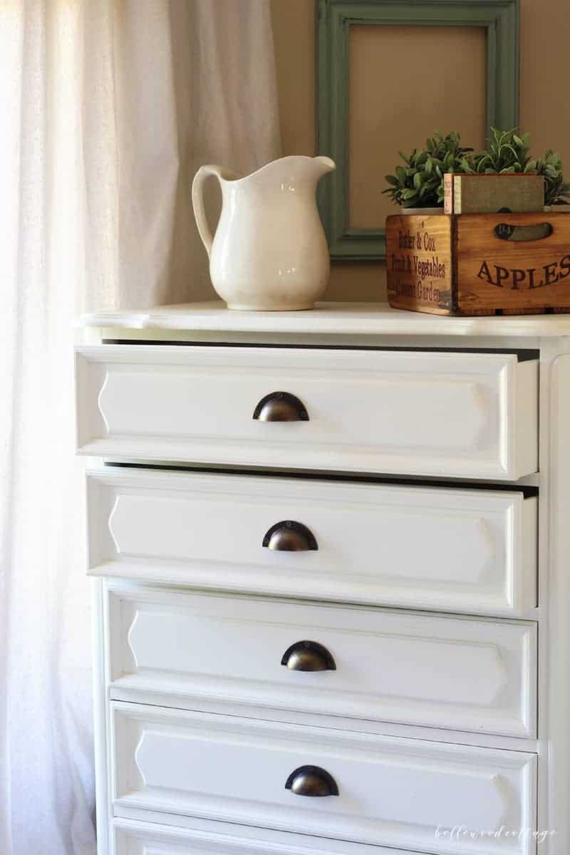 White chalk painted French country dresser with bronze cup pulls, a white ironstone pitcher and a wooden apples crate on top