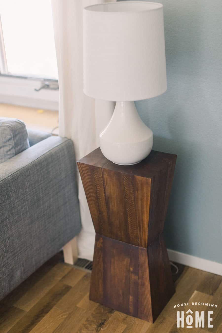 Dark stained geometric side table built from stacked tapered wood blocks beside a gray sofa, topped with a white lamp