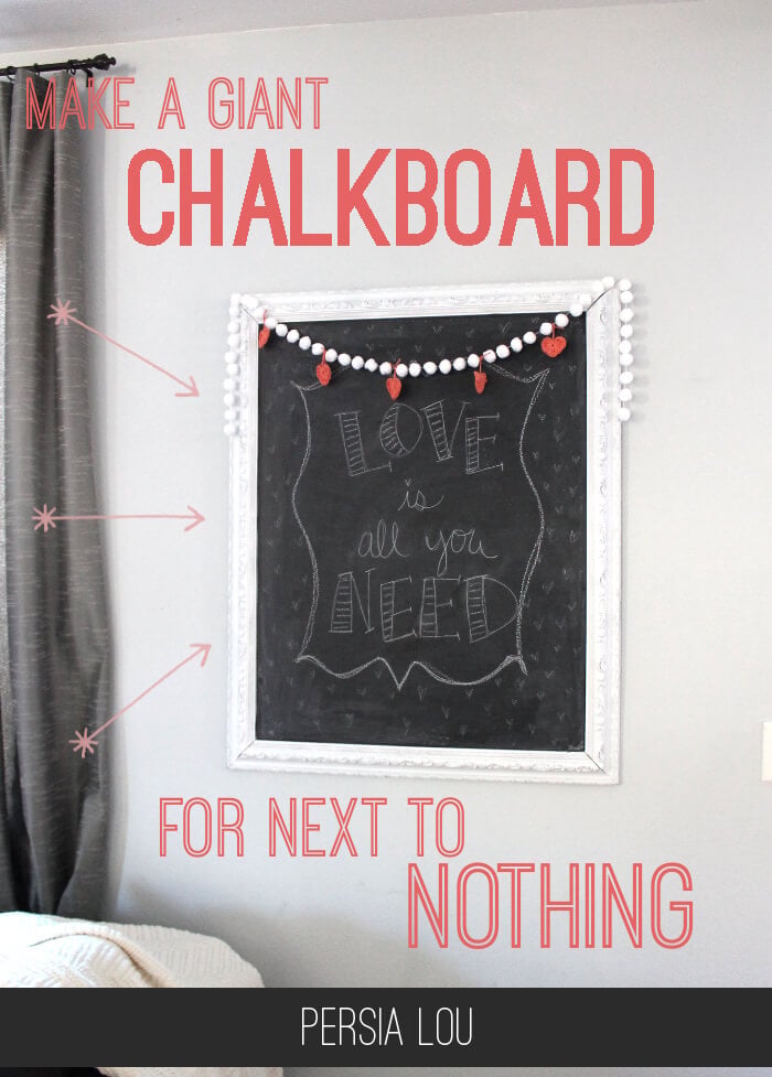 Large white framed chalkboard with hand-drawn Love is all you need lettering and a pom-pom heart garland on a gray wall
