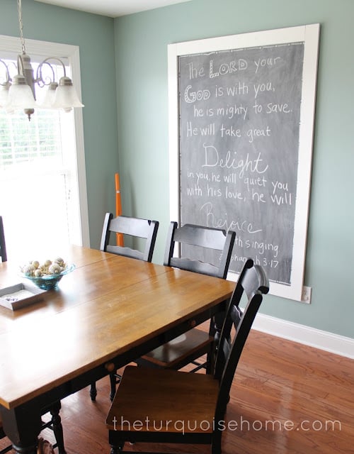 Huge framed chalkboard with chalk writing on a sage green dining room wall above a wood table with black spindle chairs