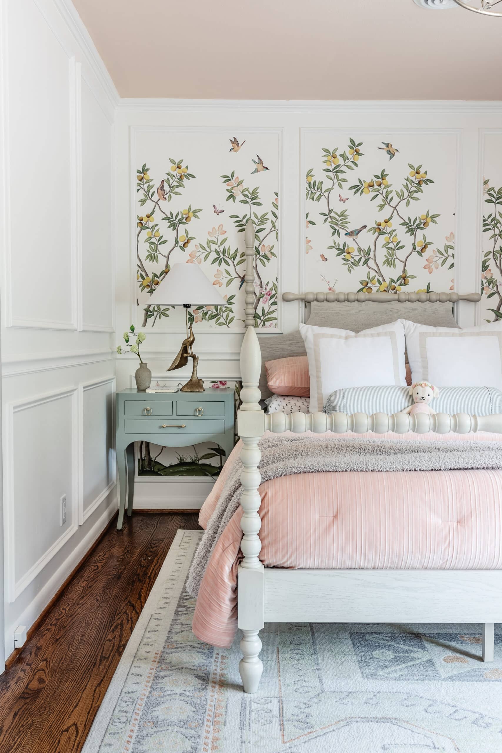 Pale aqua gloss painted bedside table with a brass lamp beside a white spindle bed and pink bedding in a floral bedroom