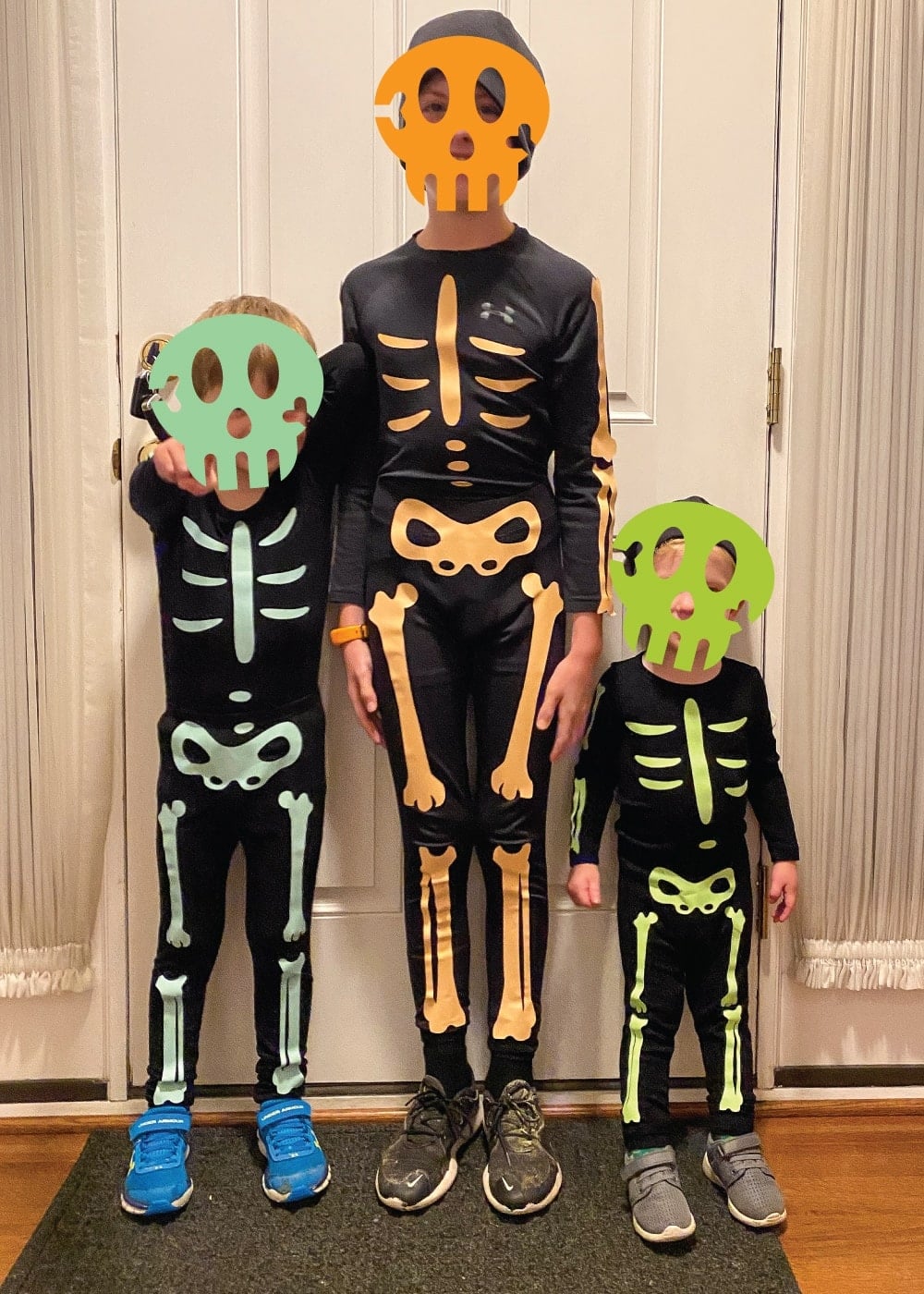 Three kids in black skeleton costumes with glow-in-the-dark vinyl bones and cut-out skull masks at a front door